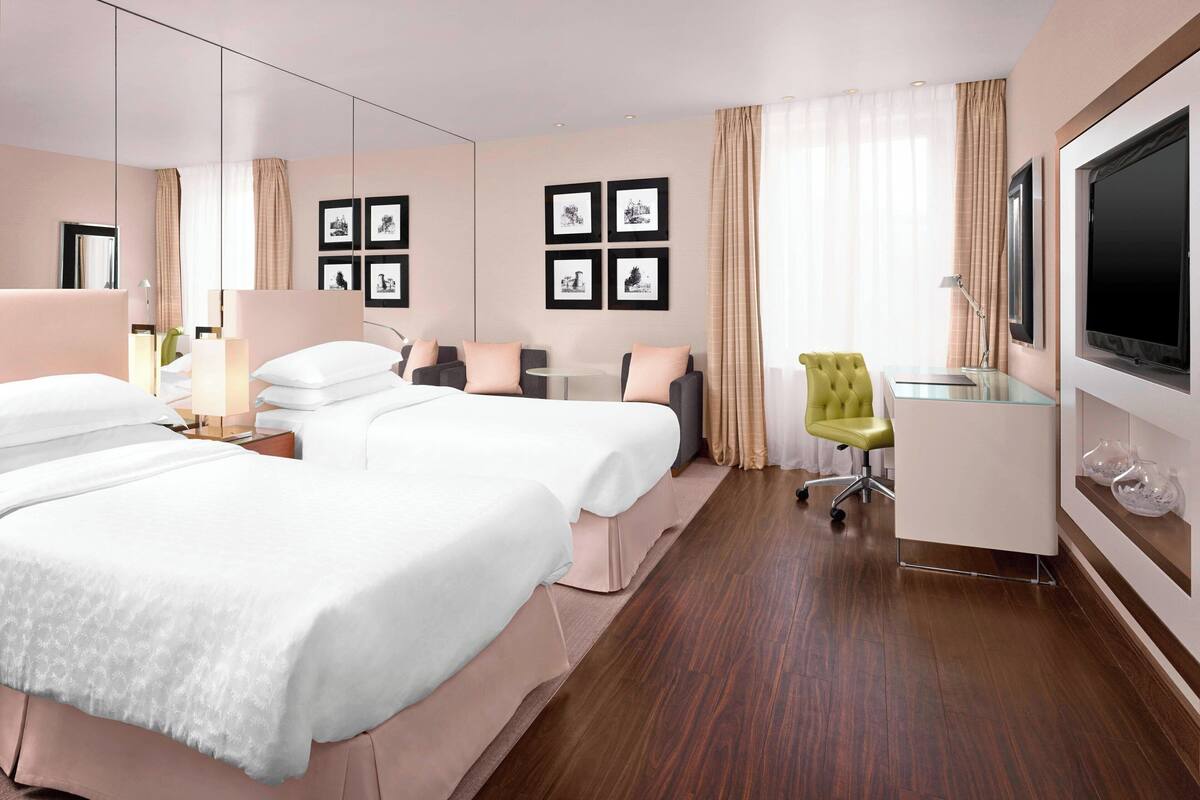 A hotel room with two beds, a desk, a chair, a TV, and framed pictures on the wall.