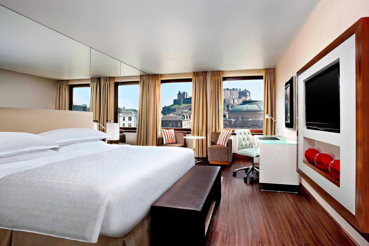 A hotel room with a large bed, a desk, a chair, a TV, and a view of a castle.