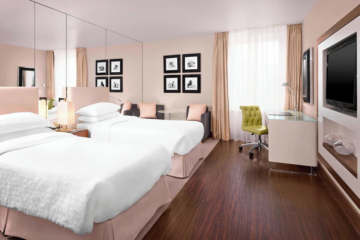A hotel room with two beds, a desk, a chair, a TV, and framed pictures on the wall.