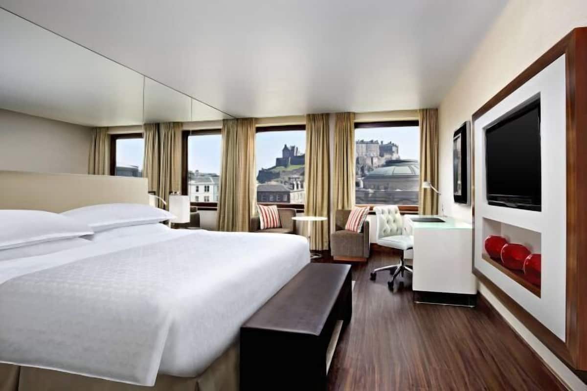 A hotel room with a large bed, a desk, a chair, a TV, and a view of a castle.