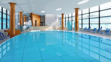 An indoor swimming pool with clear blue water, surrounded by blue lounge chairs and large windows.