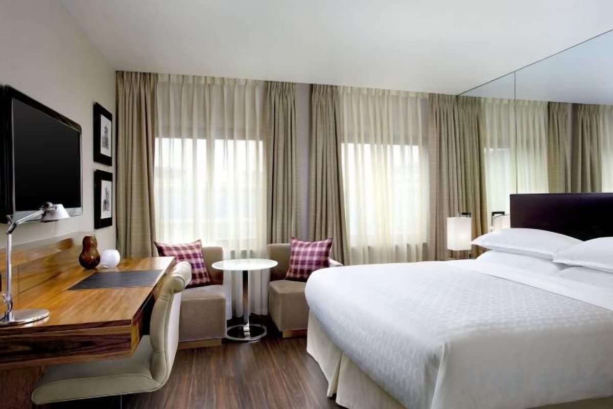 A hotel room with a bed, a desk, a chair, a small table, and a TV.