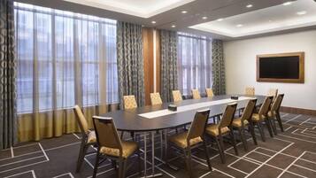 A conference room with a long table, chairs, a projector screen, and large windows with curtains.