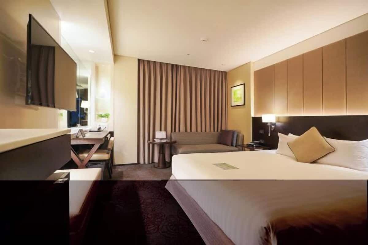 A hotel room with a bed, a desk, a chair, a small table, a lamp, and a TV.