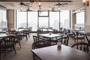 Un comedor con mesas y sillas, ventanas amplias con vista a la ciudad y ventiladores de techo.