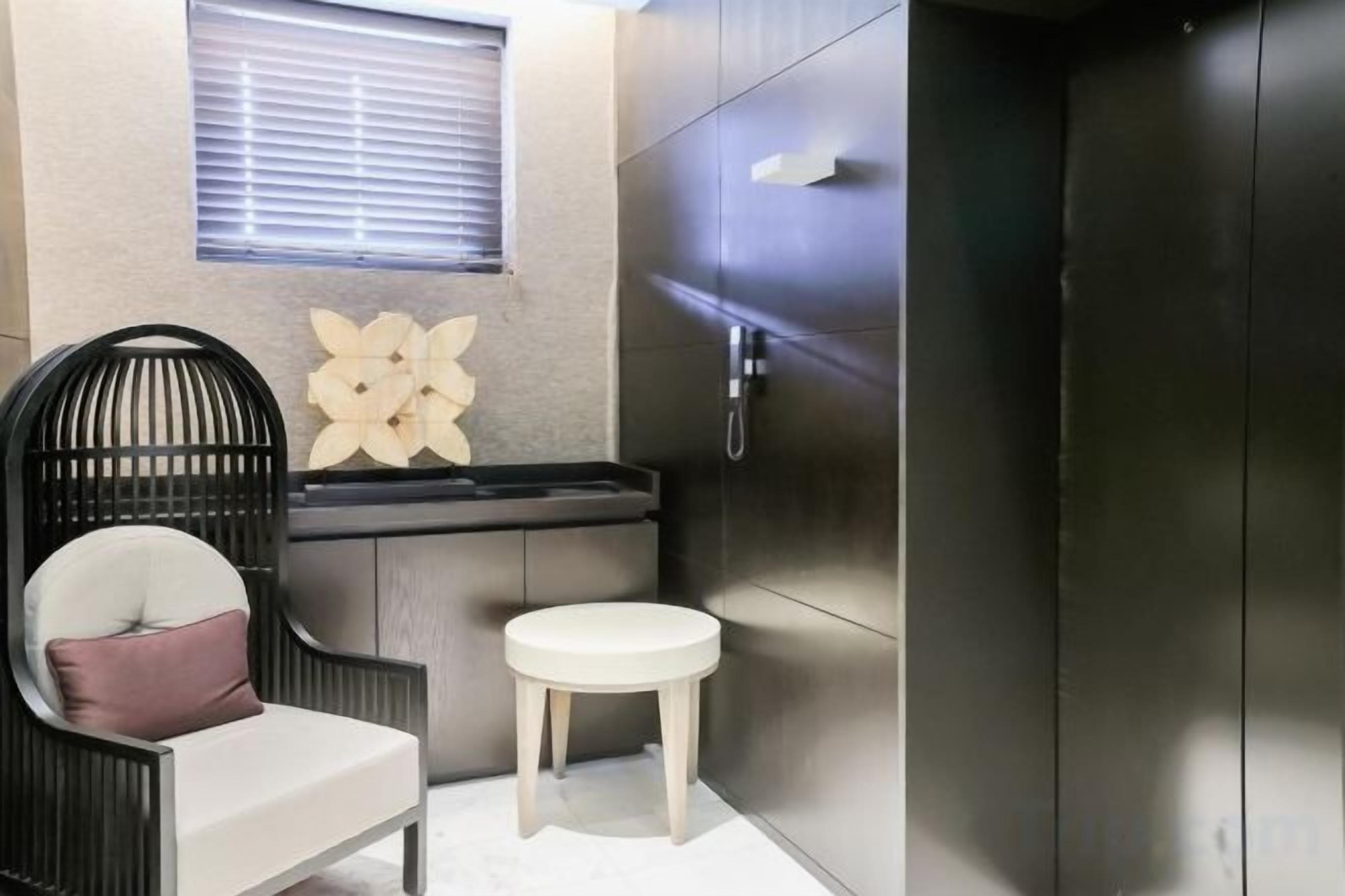 A bathroom with a black chair, a small stool, a wall-mounted sink, and a cabinet.