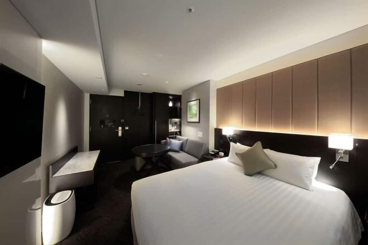A hotel room with a large bed, a desk, a chair, a TV, and a wall-mounted lamp.
