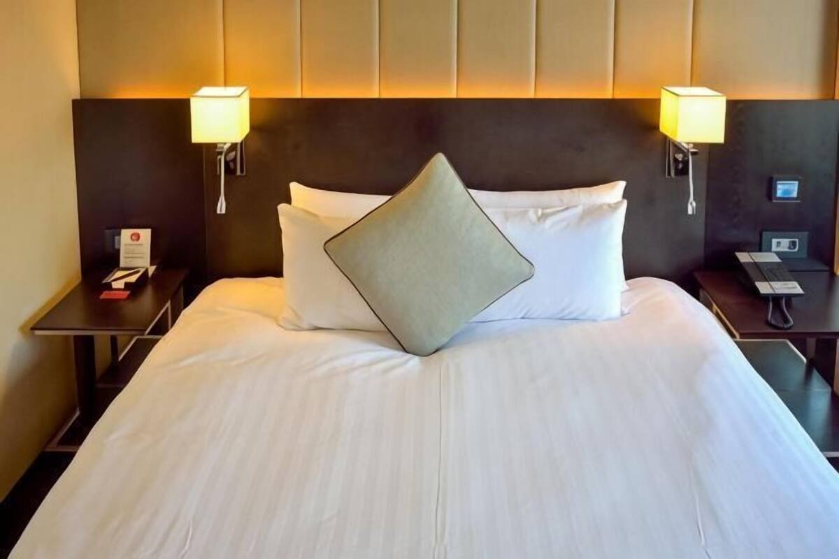 Une chambre d’hôtel avec un grand lit, deux lampes de chevet, une table de chevet avec un téléphone et un panneau de commande fixé au mur.