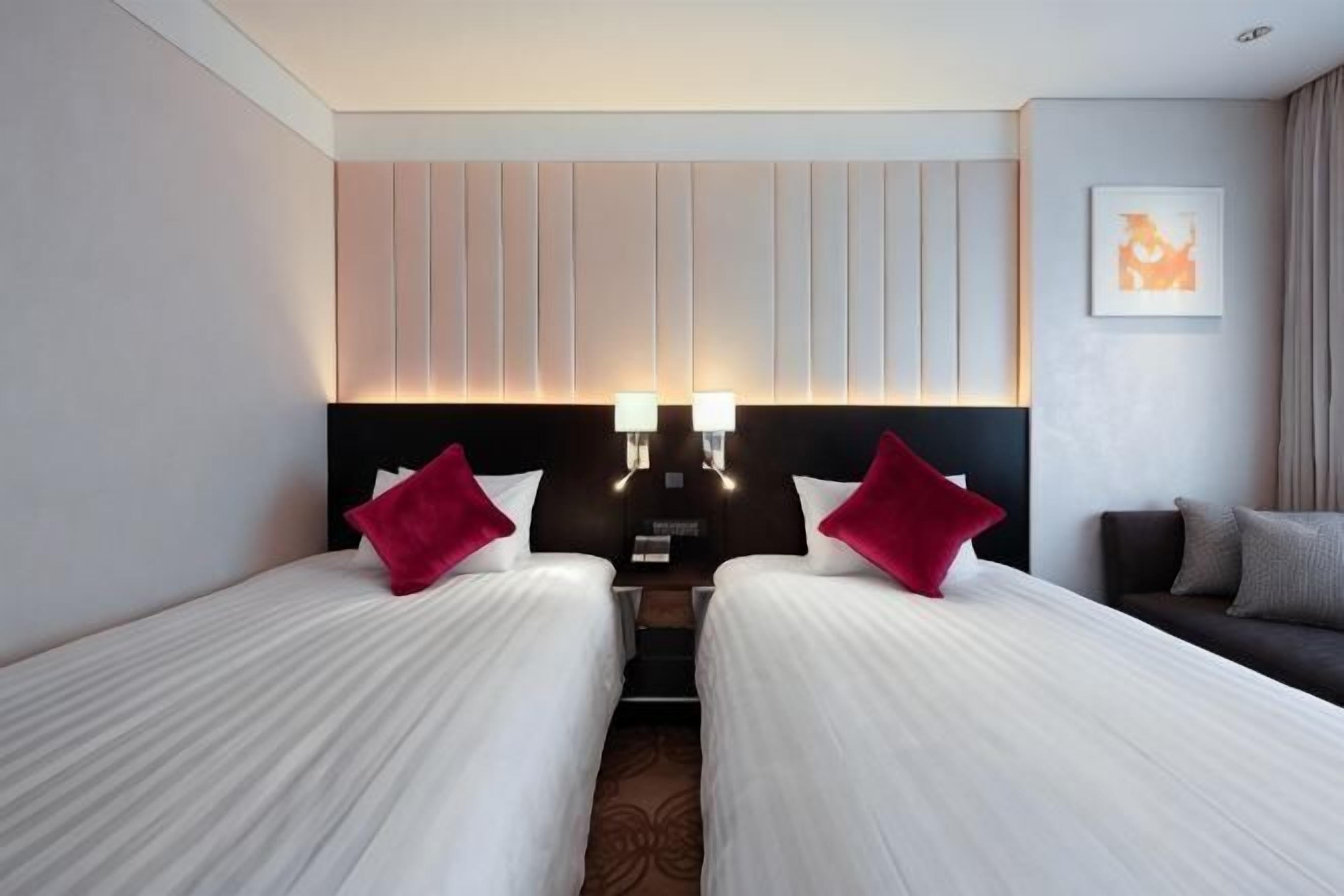 Ein Hotelzimmer mit zwei Betten, einem Kopfteil mit vertikalen Paneelen, einem Nachttisch und einer Wandlampe.