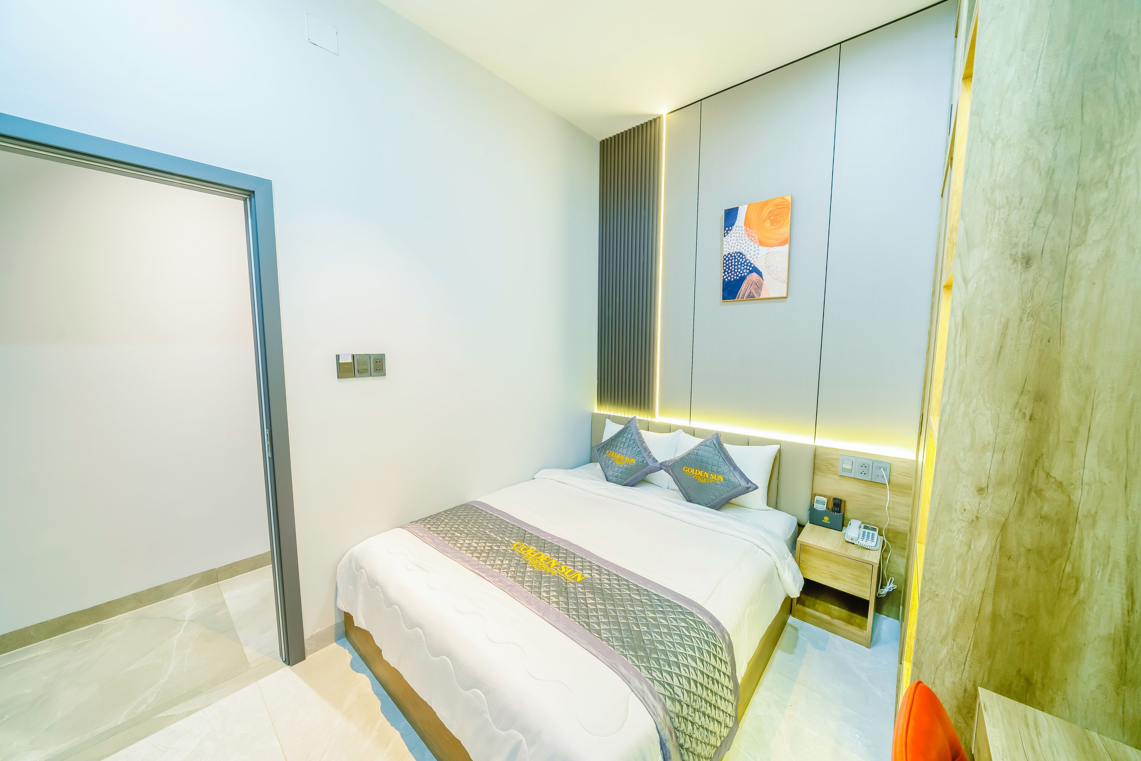 A modern hotel room with a neatly made bed, a bedside table with a phone, and a wall-mounted light fixture.