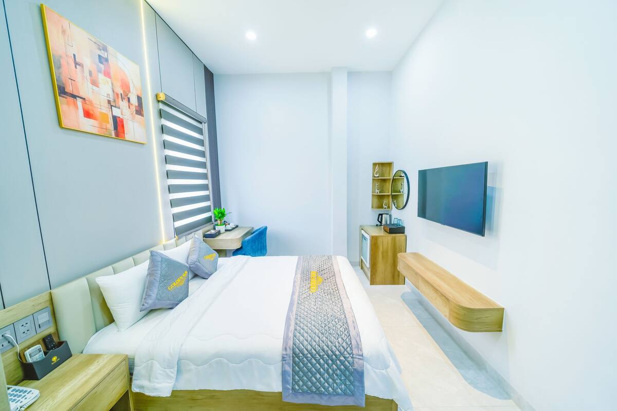 Una camera d'albergo moderna con un letto rifatto, un comodino con una lampada, una TV a parete e un piccolo armadietto in legno.