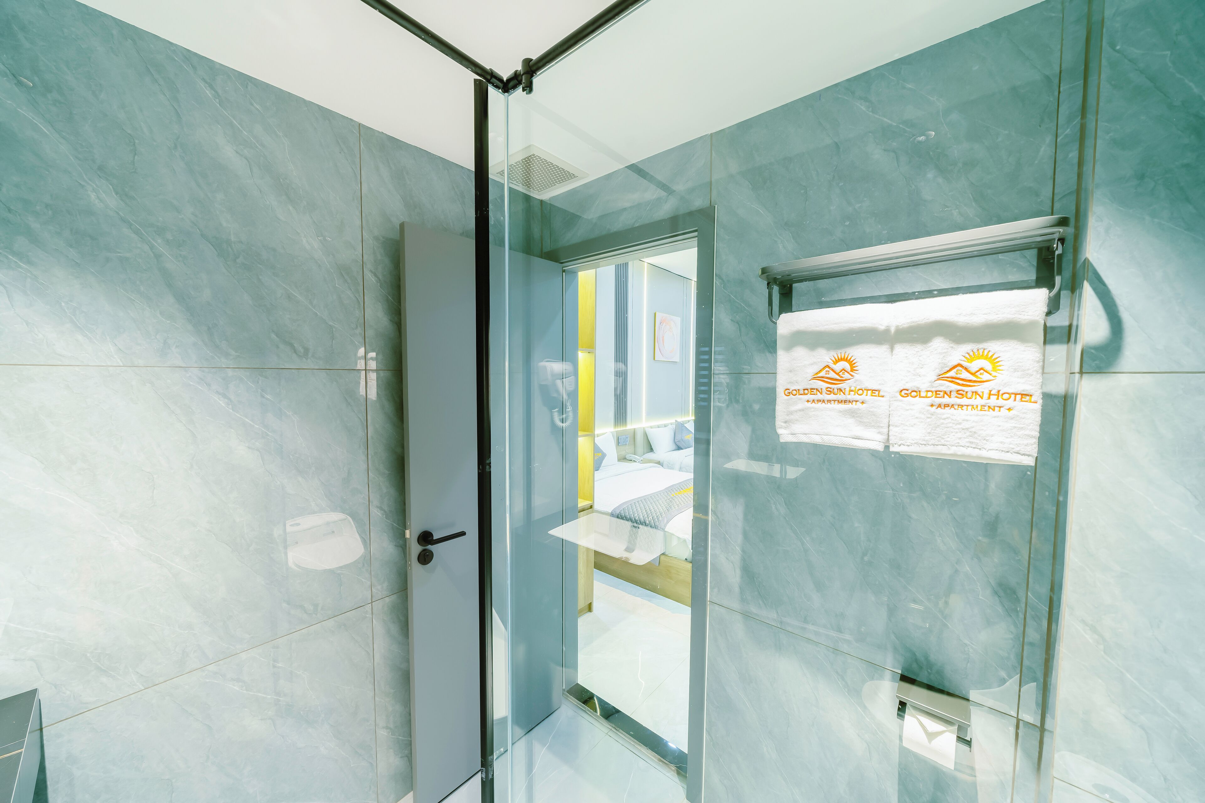 A modern hotel room with a glass door leading to a bathroom, a bed with a headboard, and a window with a view.