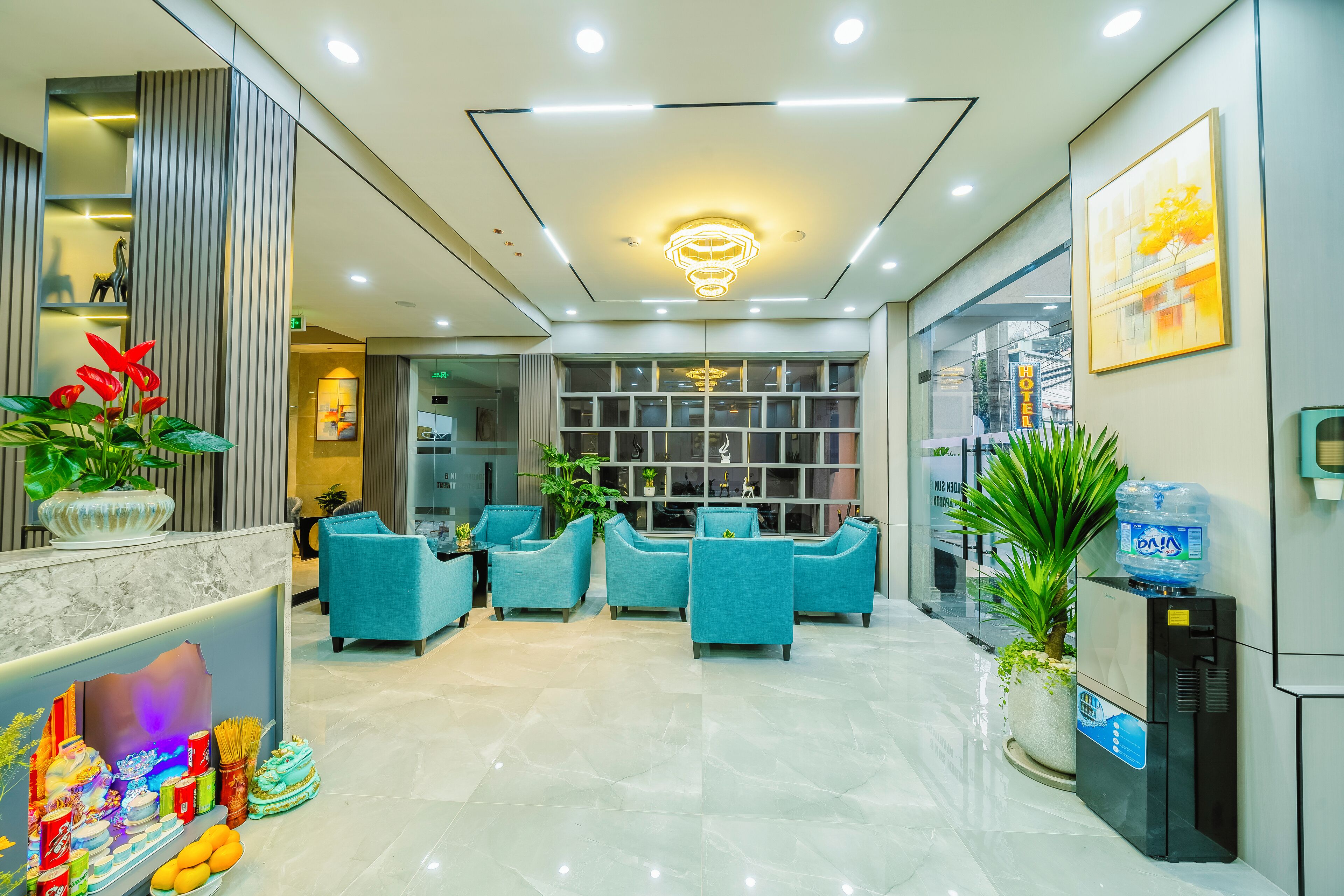 A modern hotel lobby with a marble reception desk, teal armchairs, a water dispenser, and a large window offering a view of the city.