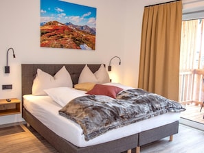 A bedroom with a large bed, a headboard, two wall-mounted lamps, a nightstand, and a window with curtains.