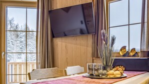 Een houten cabinteinterieur met een eettafel, stoelen, een aan de muur bevestigde televisie en zicht op besneeuwde bomen door grote ramen.