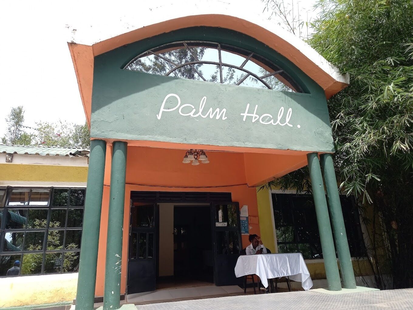 A building with a sign reading "Palm Hall." It features a curved entrance with a green and orange facade, a large window above the door, and a table with a white tablecloth outside.