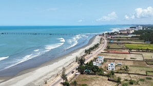 A coastal area with a beach, a pier, and buildings along the shore. - Andromeda Gazirtek (Cox's Bazar)