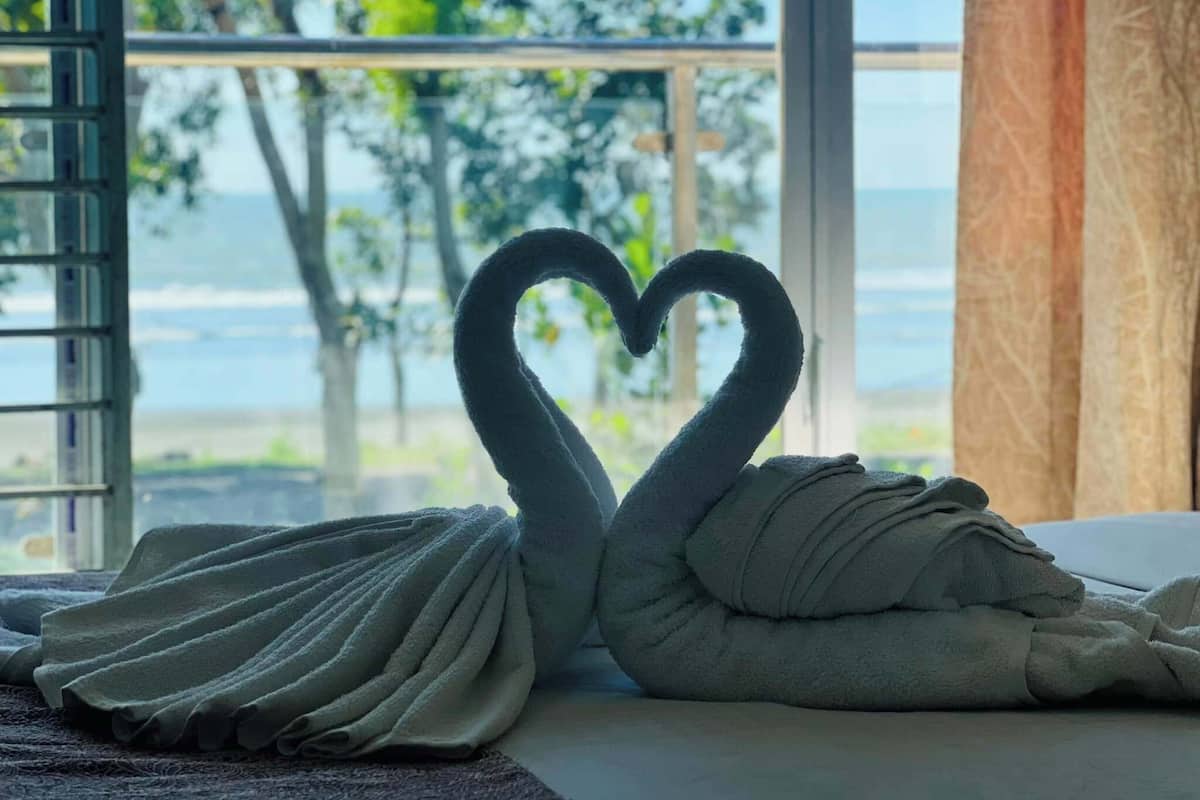 A room with a bed, a window with a view of the ocean, and a towel arranged in the shape of a heart.