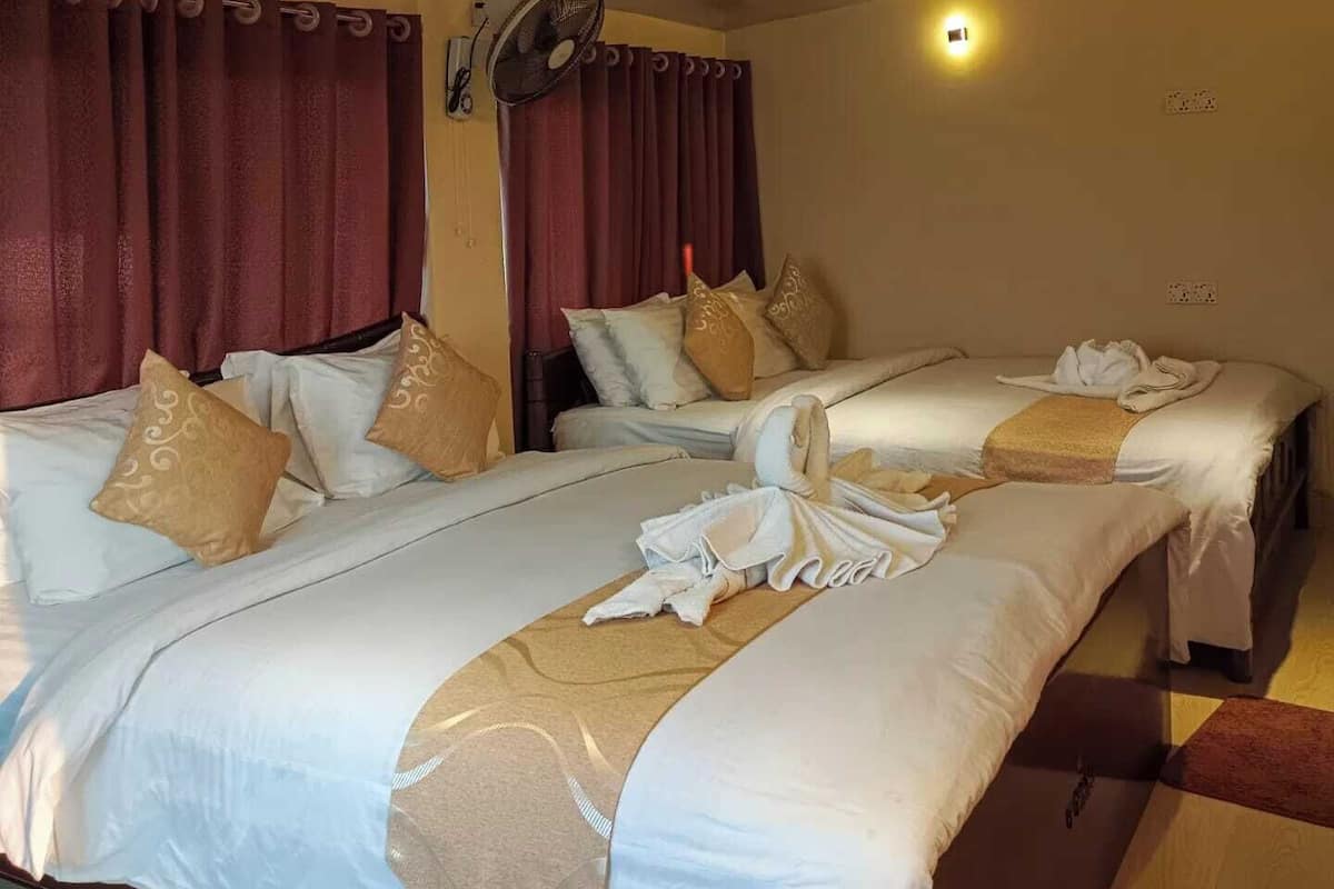 A hotel room with two beds, each with white linens and decorative pillows. There is a standing fan on the wall and a small table with a towel.