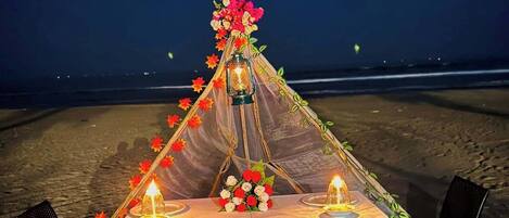 A beachside dining setup with a table set for two, adorned with candles and flowers, under a tent.