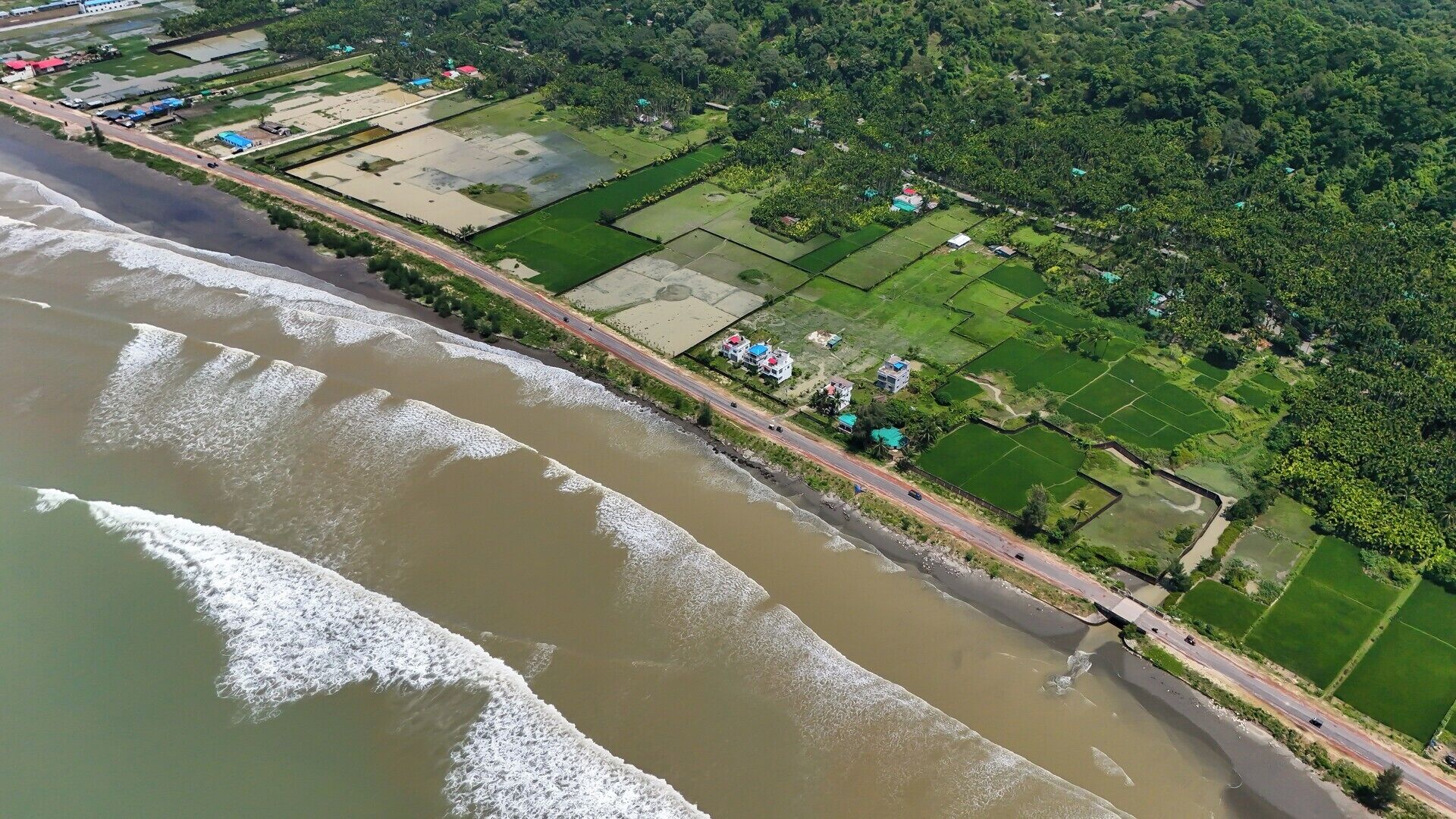 An aerial view of a coastal area with a road running parallel to the beach, green fields, and scattered buildings.