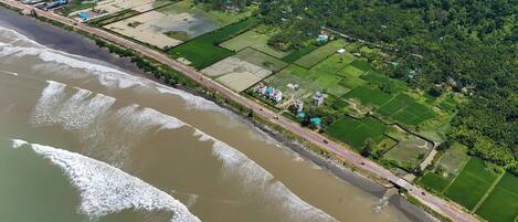An aerial view of a coastal area with a road running parallel to the beach, green fields, and scattered buildings.