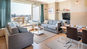 A modern living room with a grey sofa, a coffee table, and a dining area with chairs and a TV.