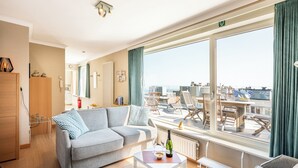 A modern hotel room with a large window, a sofa, a small table with a bottle and glasses, and a view of the cityscape.