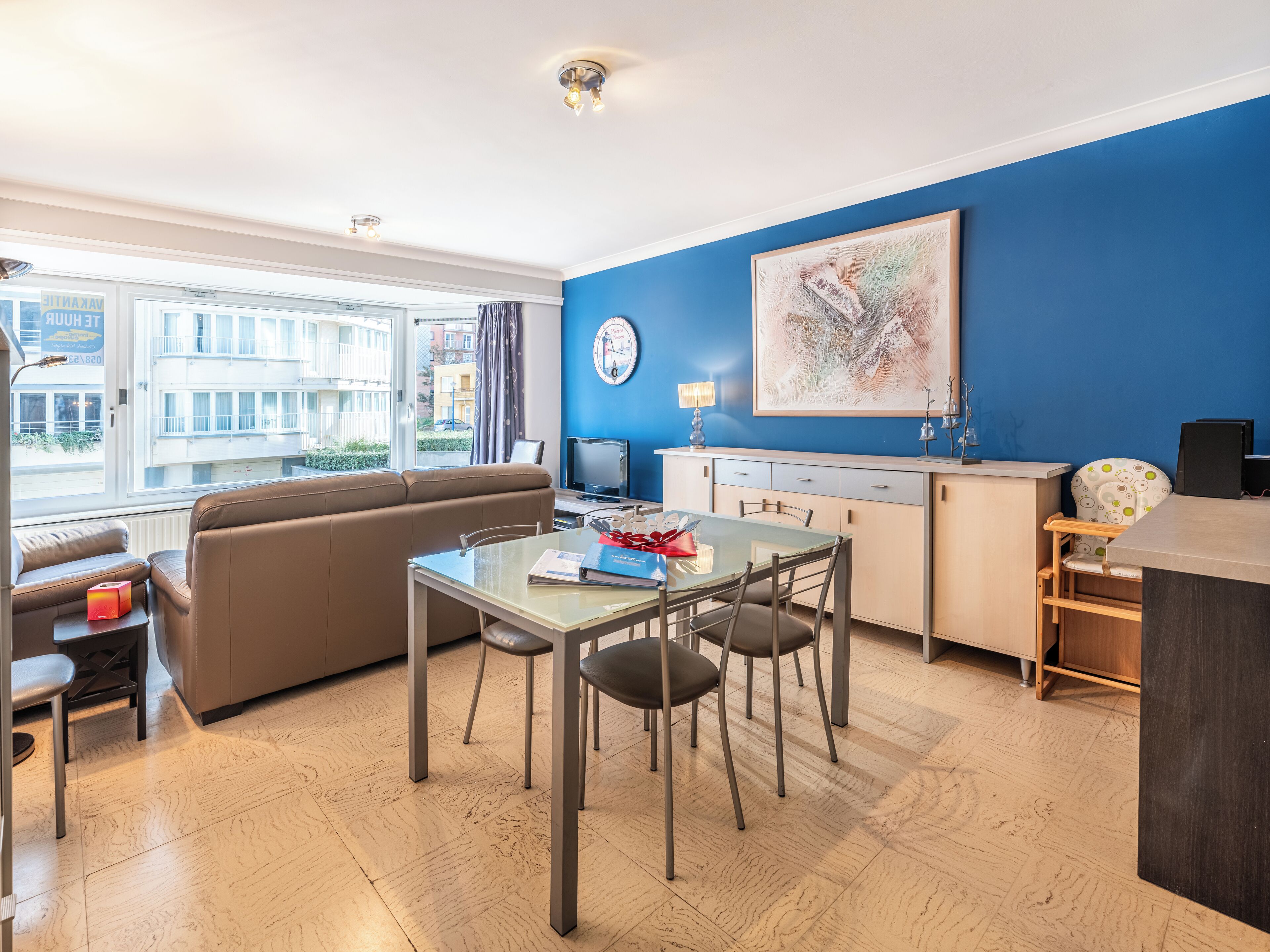 Een moderne woonkamer met een bank, een eettafel met stoelen, een keuken met kastjes en een groot raam met uitzicht op gebouwen.