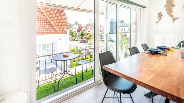 A modern dining area with a wooden table, black chairs, and a view of a residential street through large glass doors. - Renovated flat centrally located (Koksijde)