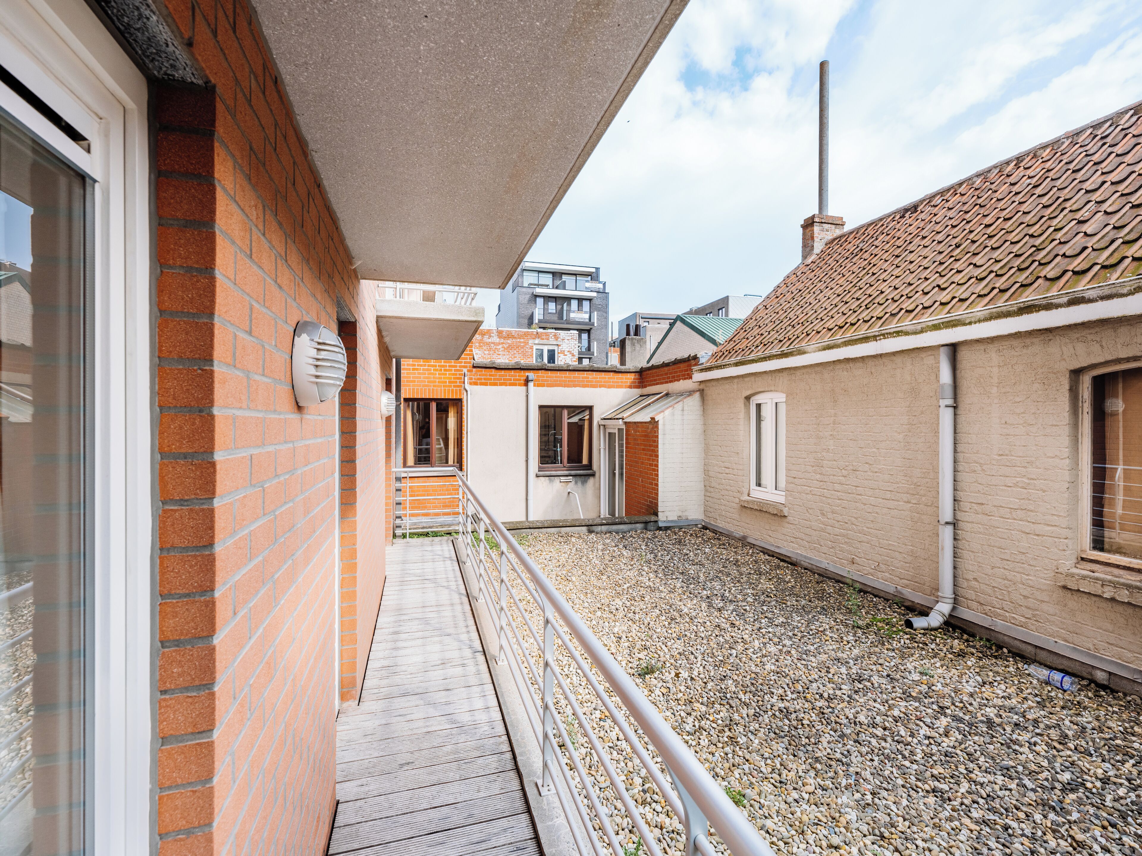 A residential building with a brick facade, a balcony with a metal railing, and a gravel courtyard.
