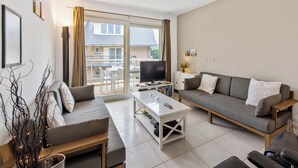 A modern living room with a sofa, a coffee table, and a TV. There is a balcony with a table and chairs outside.