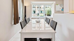 A modern dining area with a long table, chairs, and a view of a residential building through large windows.