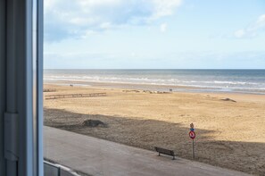 Vista a la playa desde una ventana, mostrando una playa arenosa, un banco de madera y una carretera con señal de prohibido estacionar.