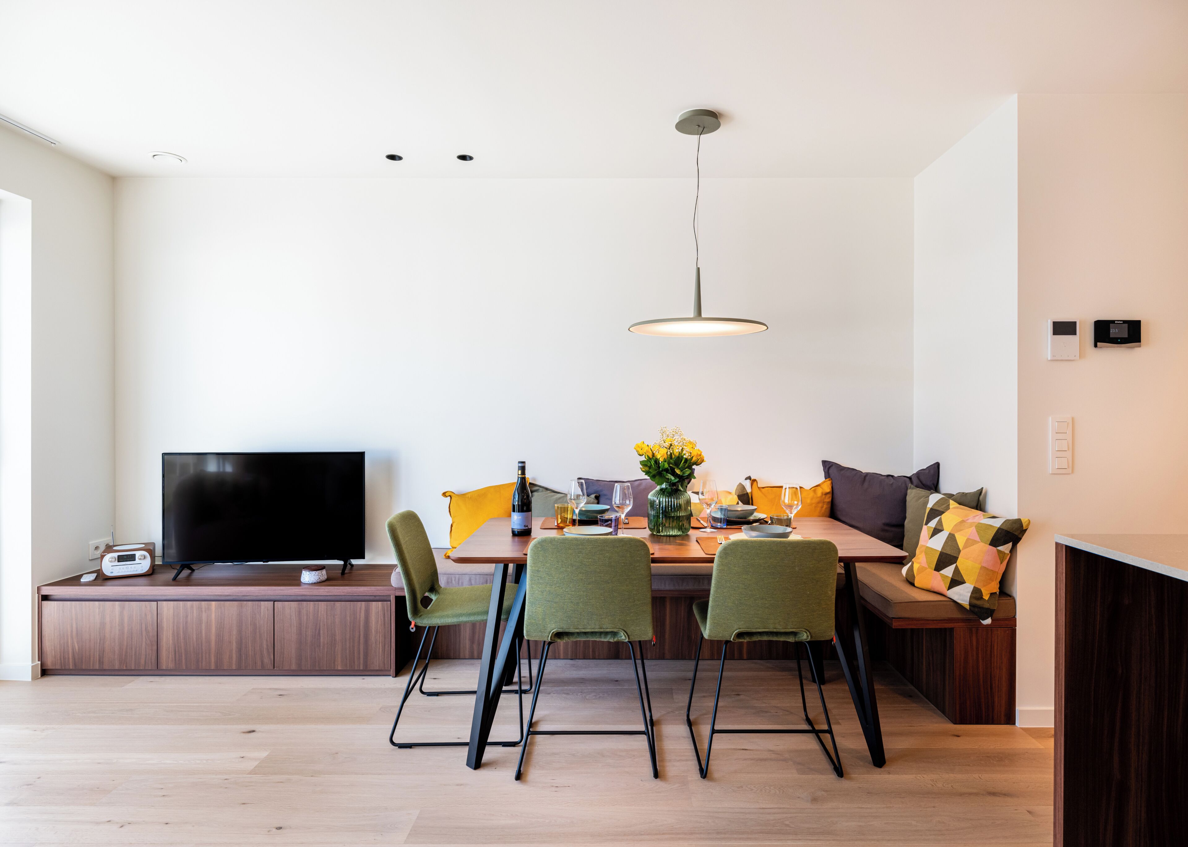 Ein modernes Wohnzimmer mit Flachbildfernseher, einem Holzesstisch, grünen Stühlen und einer gelb gepolsterten Couch.