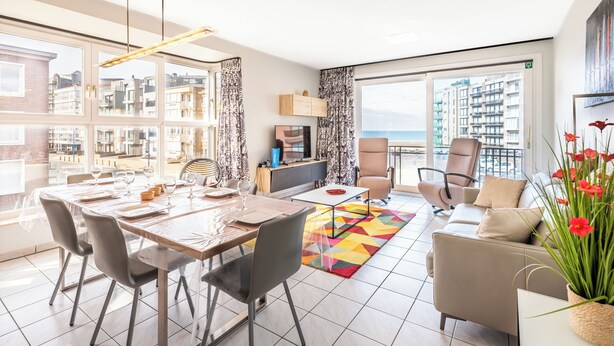 A modern living room with a dining area, large windows, and a view of the cityscape. - Beautifully renovated flat with sea views (Koksijde)