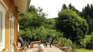 Una terraza de madera con mobiliario exterior, una jardinera de piedra y un jardín con diversas plantas y árboles.