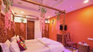 A bedroom with a wooden bed, hanging pink flowers, a wooden chair, and a small round table with a lamp. - Neev Haveli (Udaipur)
