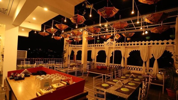 An indoor dining area with multiple tables and chairs, decorative hanging lamps, and a buffet table with food items. - Neev Haveli (Udaipur)