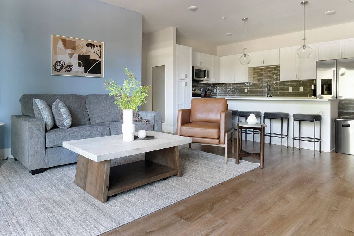 A modern living room with a grey sofa, a coffee table, and a brown leather chair. A kitchen with white cabinets and a stainless steel refrigerator is visible in the background.