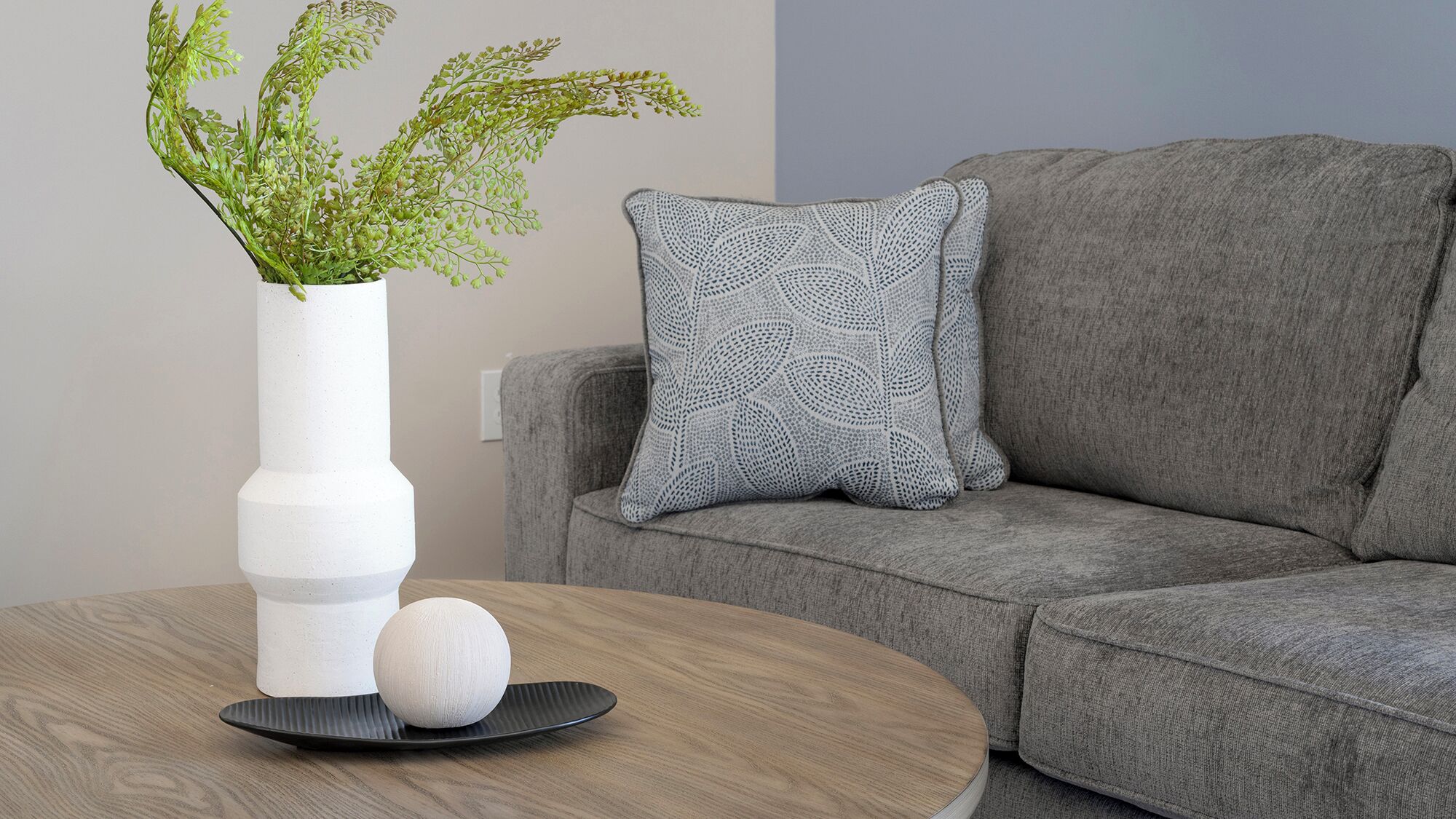 Un salon moderne avec un canapé gris, un vase blanc orné de feuillage vert, un coussin gris à motifs, une table ronde en bois et un objet sphérique blanc.