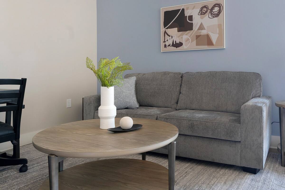 A modern living room with a grey sofa, a round wooden coffee table, a white vase with green plants, a black chair, and a lamp on a side table.