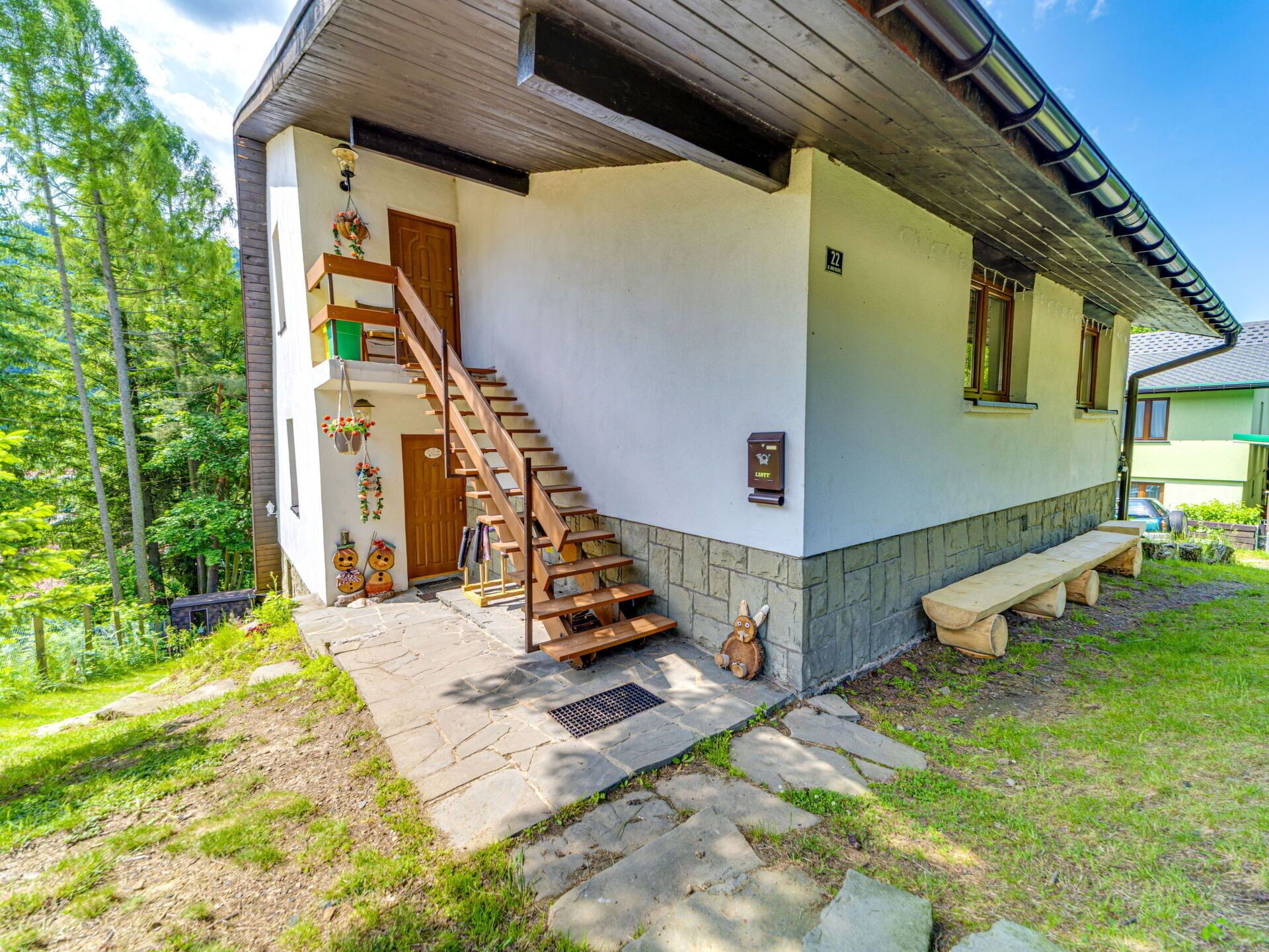 A two-story house with a wooden staircase leading to a front door, a bench outside, and a stone pathway.