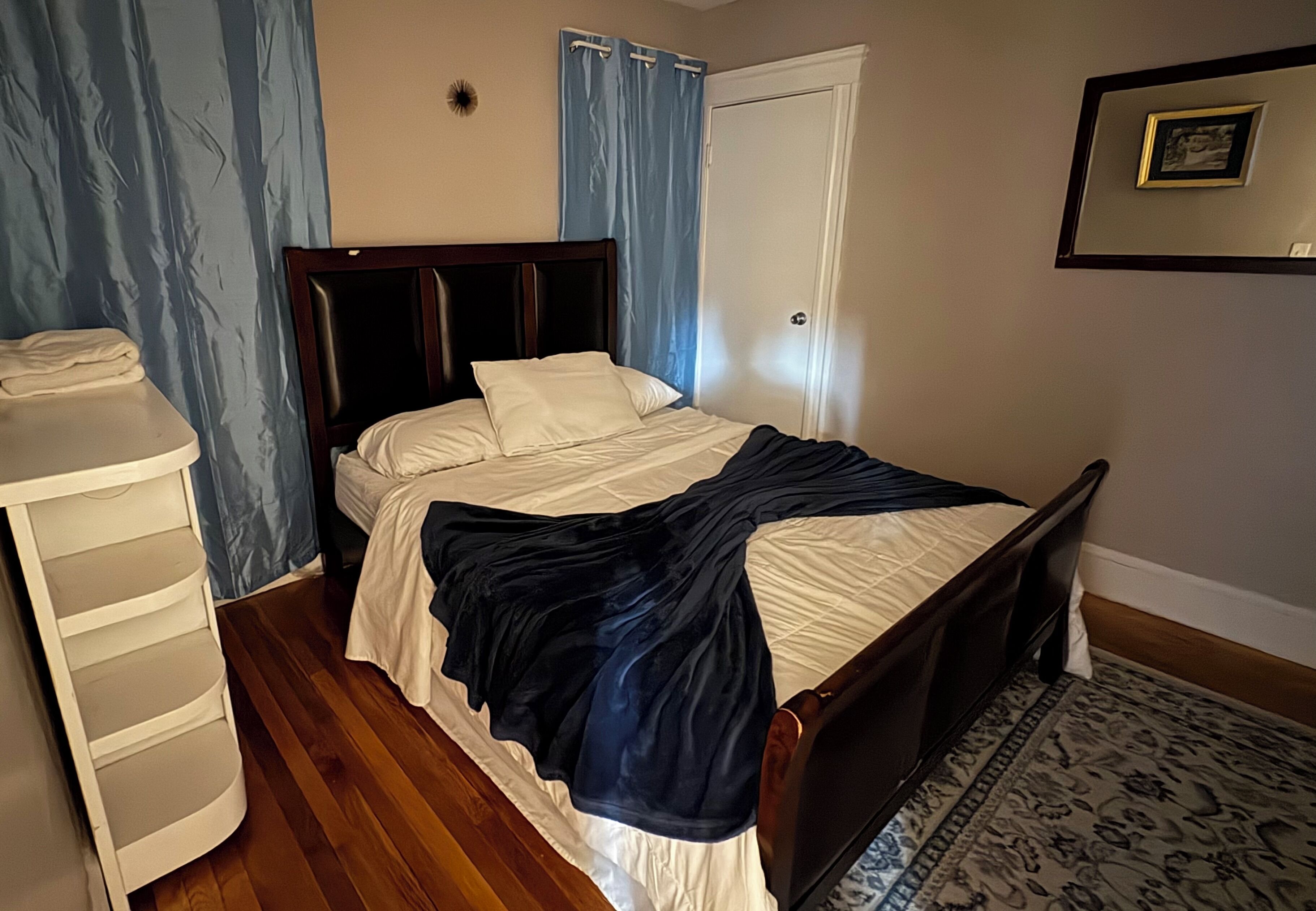 A bedroom with a wooden bed, a white dresser, a blue curtain, and a framed picture on the wall.
