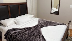 A neatly made bed with white linens and a dark grey blanket, a wooden headboard, a mirror on the wall, and a light-colored curtain. - Cozy large 2 bedrooms apartment on Historic Mt Auburn • 5 Min to Harvard (Watertown)