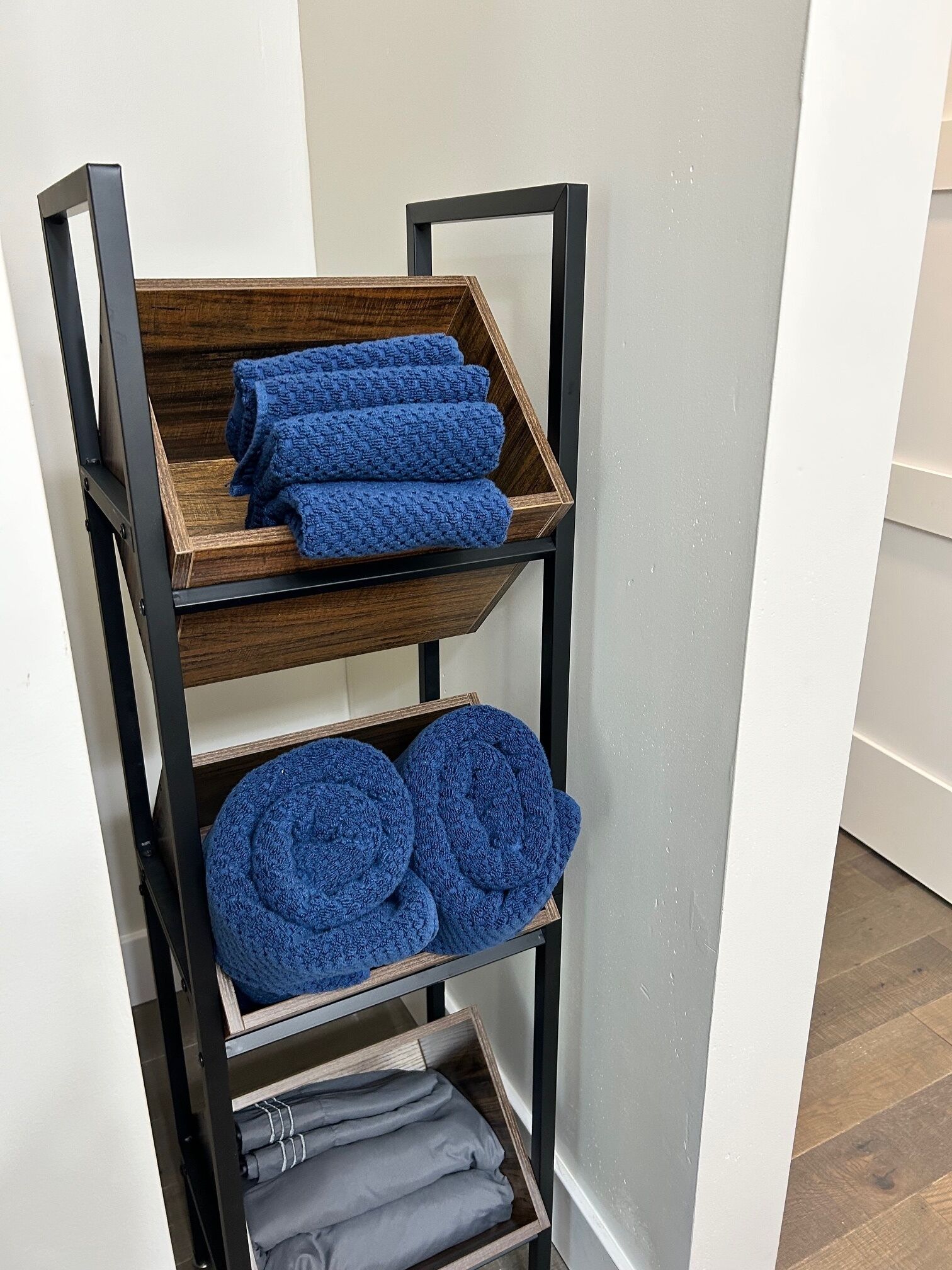 A wooden shelf unit with three tiers, each holding rolled towels in blue and gray colors.