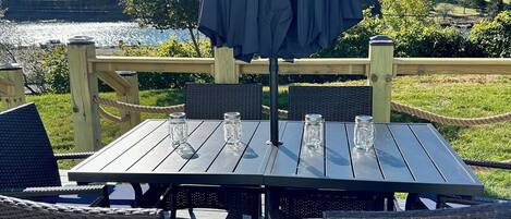 Eine Terrasse mit Tisch und Stühlen, einem schattenspendenden Sonnenschirm und Blick auf See und Bäume.