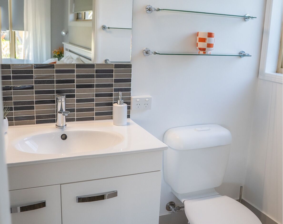 Een badkamer met een witte wastafel, een toilet en een spiegel boven de wastafel. Boven het toilet zijn er twee planken.
