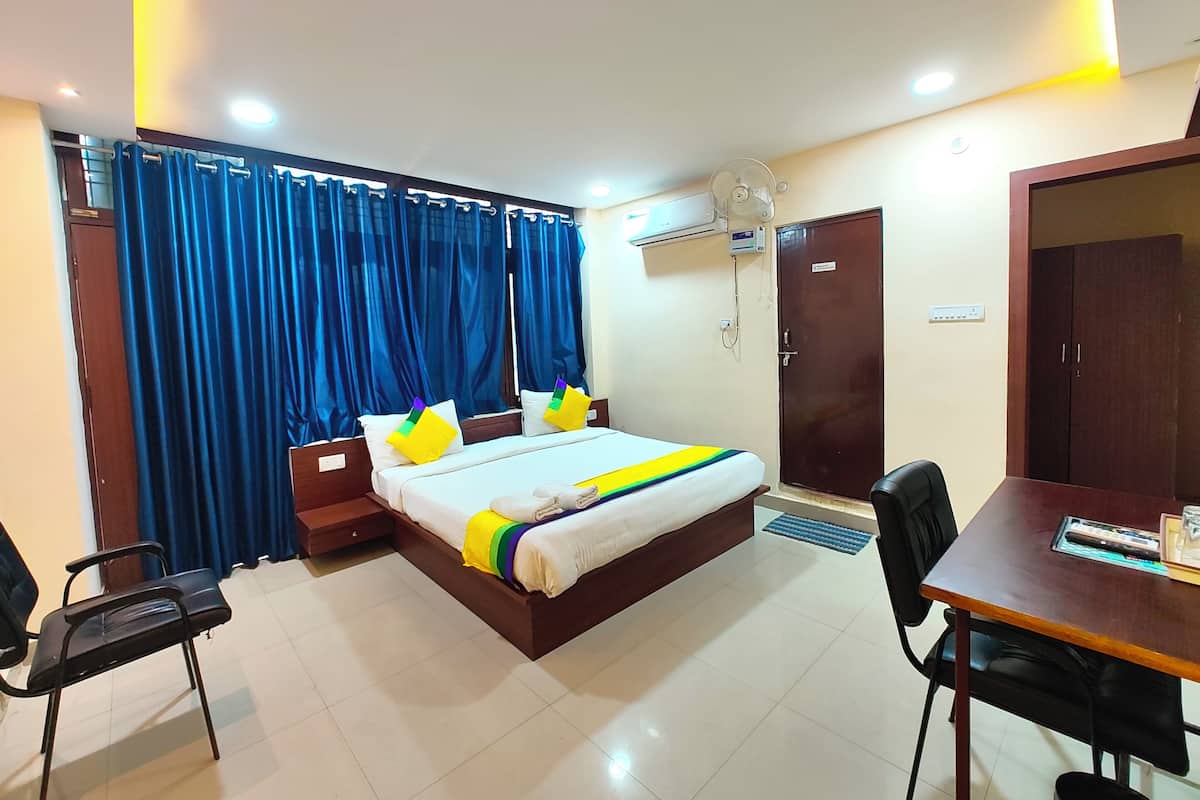 A hotel room with a bed, a desk, and a chair. There is a window with blue curtains, an air conditioning unit, and a door.