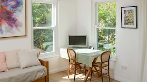 A cozy room with a sofa, a dining table with chairs, a TV, and large windows offering a view of greenery.